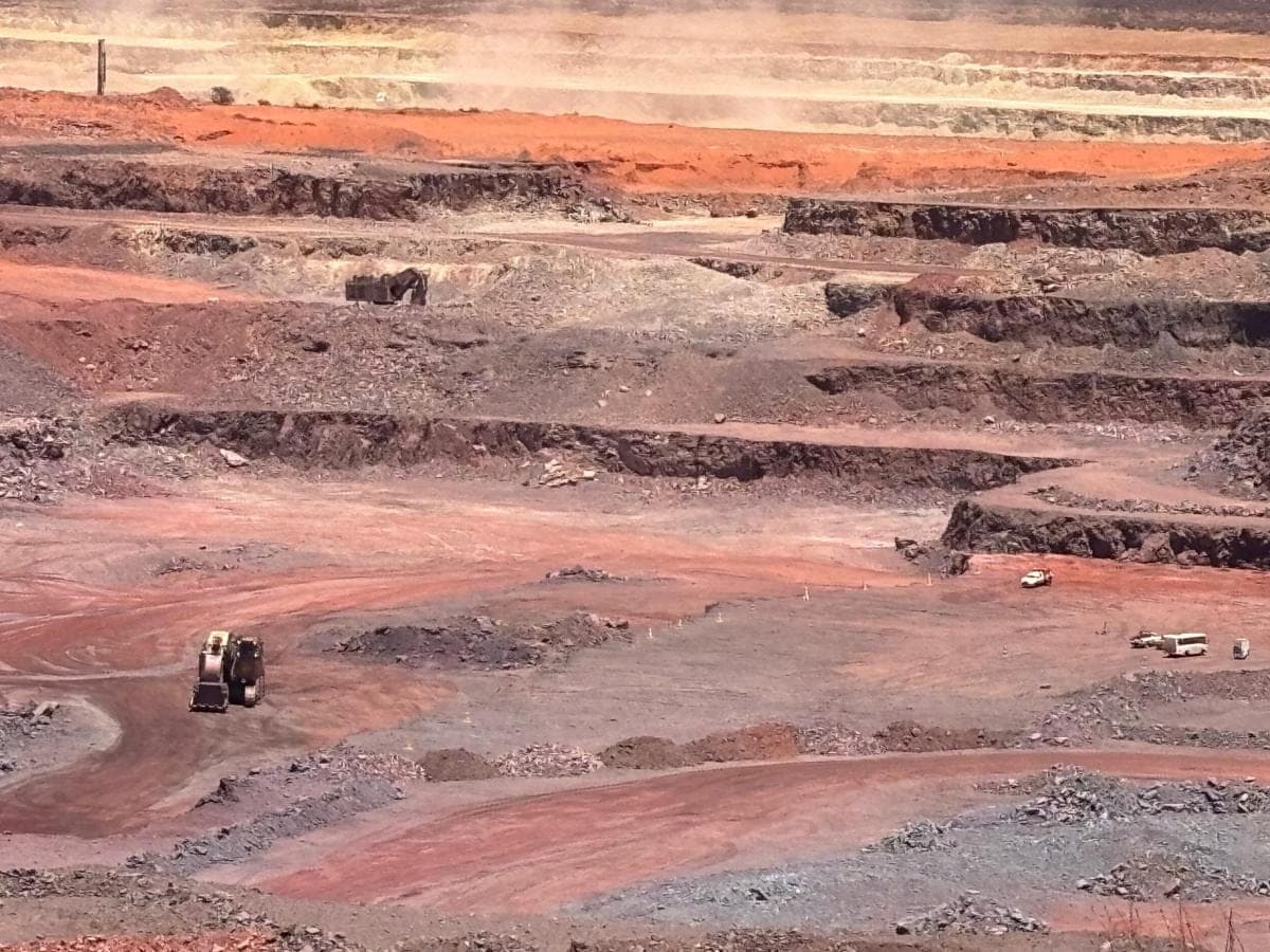 Terraced benches of a red iron-ore open pit, with an excavator and a front-end loader working the floor.