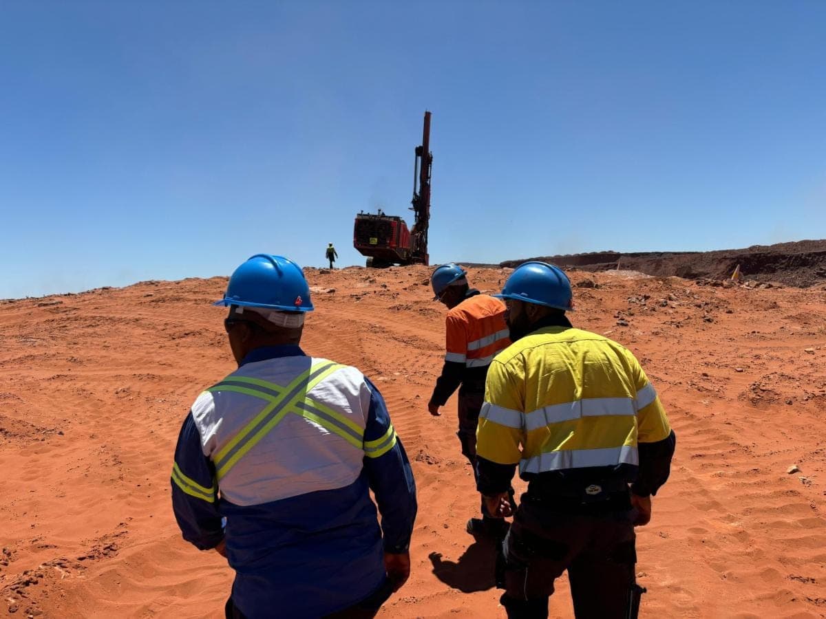 Three site personnel in hard hats and reflective wear walking toward a surface drill rig on a red-sand bench.