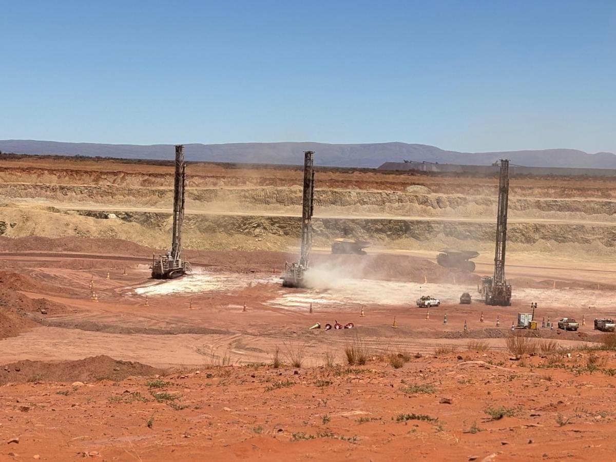 Three surface drill rigs collaring a blast pattern on a bench, with haul trucks working behind them.