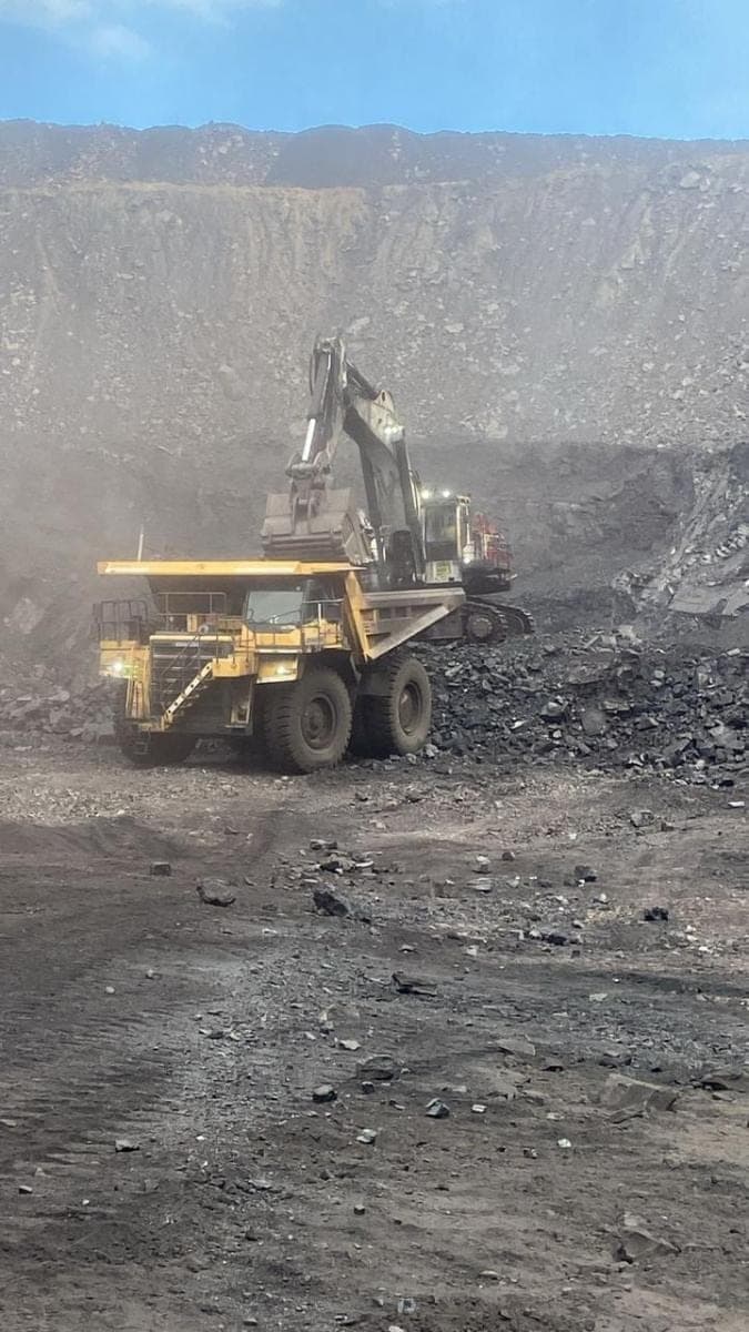 An excavator swinging a loaded bucket over a haul truck in heavy dust.