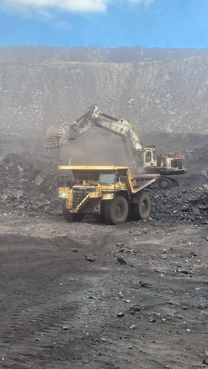 An excavator loading a haul truck from a coal face, dust hanging across the bench.