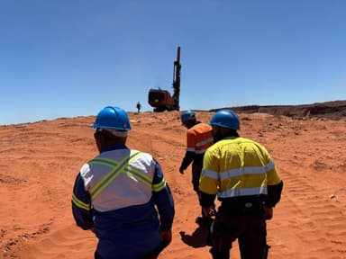 Three site personnel in hard hats and reflective wear walking toward a surface drill rig on a red-sand bench.
