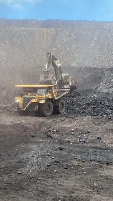 An excavator swinging a loaded bucket over a haul truck in heavy dust.