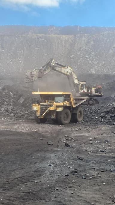 An excavator loading a haul truck from a coal face, dust hanging across the bench.